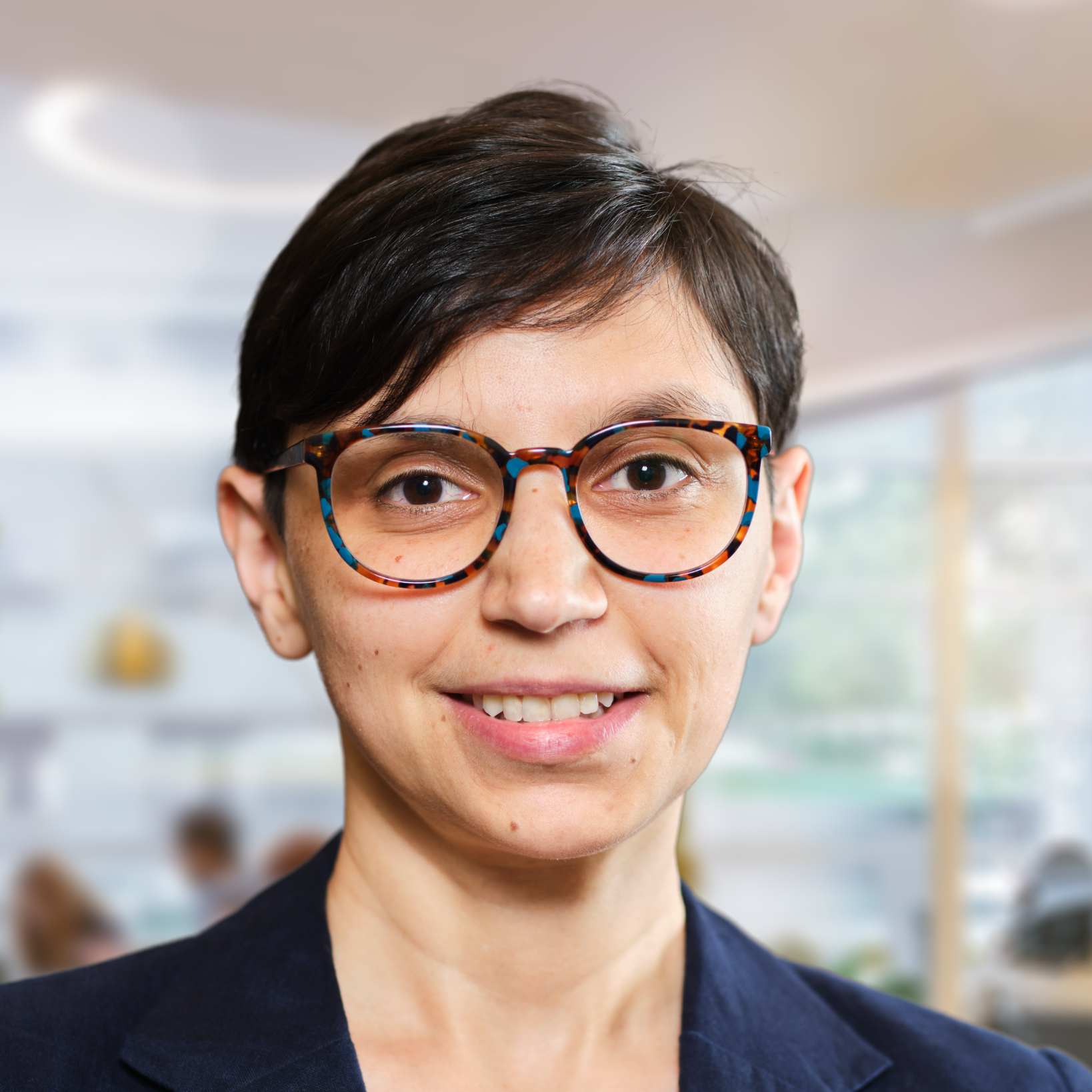 Caucasian woman with dark brown hair (in a pixie cut) and black-rimmed glasses, wearing a blazer, with a whiteboard, desk, and other office sundries behind her