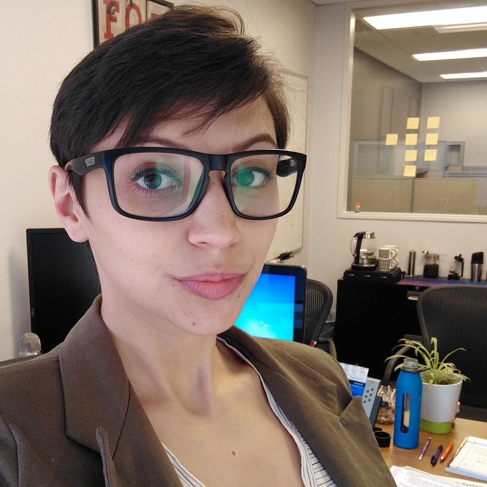 Caucasian woman with dark brown hair (in a pixie cut) and black-rimmed glasses, wearing a blazer, with a whiteboard, desk, and other office sundries behind her